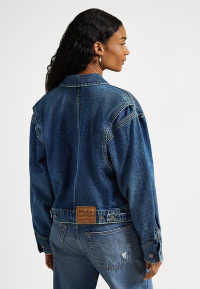 Kvinne iført en blå denimjakke med en Polo-merkelapp på baksiden, kombinert med lyseblå, slitte denimjeanser, som ser til siden.