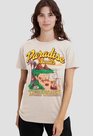 Woman wearing beige t-shirt with colorful tropical island print, palm trees, shipwreck, and text "Paradise Awaits Little Garden."