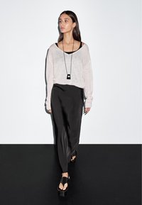 Frau geht und trägt einen lockeren hellen Pullover über einem schwarzen Oberteil, einen langen schwarzen Satinhrock, Plateausandalen und eine lange schwarze Anhängerkette.