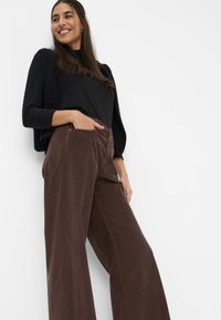 Braune Cordhosen mit weitem Bein und vorderen Taschen, kombiniert mit einem schwarzen Oberteil und einer schwarzen, strukturierten Weste, Modell steht vor einem weißen Hintergrund.