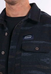 Hombre con camisa de cuadros oscuros de botones y bolsillo en el pecho que muestra la etiqueta de la marca "OXBOW" y camiseta interior negra.