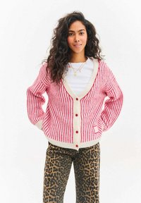 Pruhovaný růžový kardigan s krémovými akcenty a červenými knoflíky, s výstřihem do V a žebrovanou texturou. Kombinován s kalhotami s leopardím potiskem.