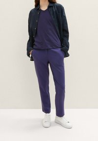 Marinblå skjorta med knappar över en lila t-shirt med v-ringning, kombinerad med lila byxor och vita sneakers. Slät tyg med avslappnad passform.