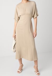 Béžové midi šaty s krátkými, ohrnutými rukávy, nařaseným živůtkem a hladkou texturou. K nim je oblečena kabelka světlé barvy a sandály na blokovém podpatku.