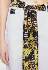 Jeans blancos de cintura alta con una bufanda de patrón barroco en negro y dorado atada como un cinturón alrededor de la cintura, y una mano visible descansando a un lado.