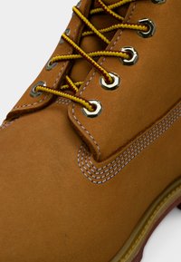 Botte en cuir marron avec des lacets jaunes et rouges passés dans des œillets argentés et une couture blanche détaillée sur le côté.
