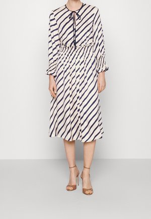 Robe à manches longues crème avec des rayures diagonales bleu marine, dotée d'un col noué et d'une taille plissée. Portée avec des talons à brides tannée.