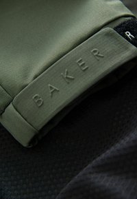 Etichetta di stoffa verde con la scritta "BAKER" in rilievo, attaccata a un materiale nero testurizzato con un motivo geometrico. Linee pulite, superficie morbida.