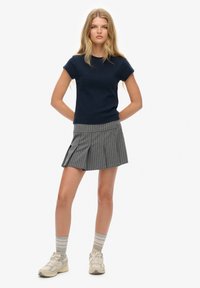Navy korte mouw gebreid shirt in combinatie met een grijs gestreepte geplooide rok. Model draagt lichte sneakers en gestreepte sokken. Minimalistisch ontwerp en kleurenpalet.
