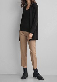 Svart lång cardigan över en svart linne, kombinerat med beigefärgade tapered-byxor och svarta snörboots på en grå bakgrund.