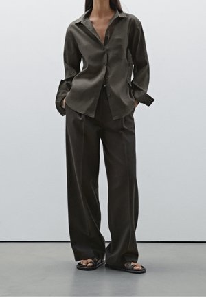 Woman wearing loose dark brown button-up shirt and wide-leg pants, standing with hands in pockets, wearing black sandals on gray floor.