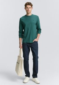 Grünes langärmliges Shirt mit rundem Halsausschnitt, kombiniert mit dunklen Jeans. Das Model hält eine beigefarbene Tragetasche mit einem Logo. Die Schuhe sind weiße Sneakers.