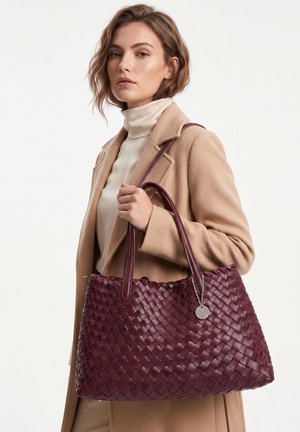Vrouw met een beige jas en coltrui die een grote bordeauxrode geweven leren draagtas met dubbele handvatten en metalen label vasthoudt.