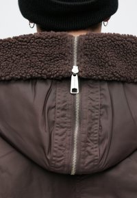 Καφέ σακάκι με μία χνουδωτή, καφέ επένδυση και ασημένιο φερμουάρ. Το υφαντό ύφασμα είναι μαλακό και λείο, με σχεδίαση χαλαρού εφαρμογής.