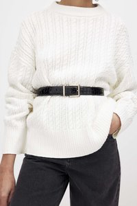 White knitted sweater with cable pattern, rounded neckline, and ribbed cuffs. Black crocodile-patterned belt at waist, paired with dark jeans.