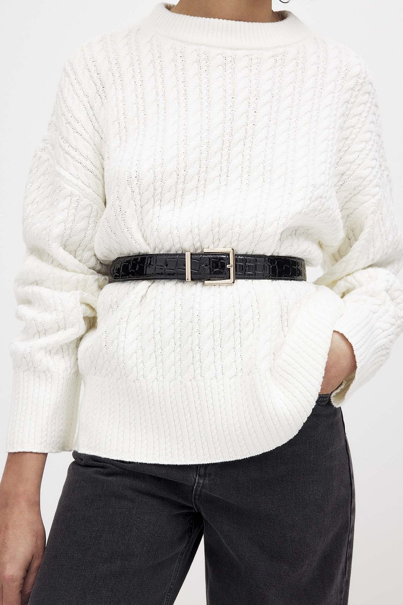 White knitted sweater with cable pattern, rounded neckline, and ribbed cuffs. Black crocodile-patterned belt at waist, paired with dark jeans.