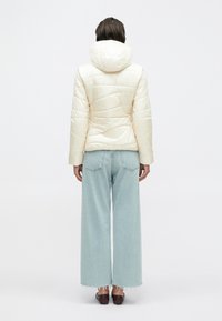 Giacca piumino color crema con design trapuntato, cappuccio e maniche lunghe, abbinata a jeans a gamba larga azzurri.