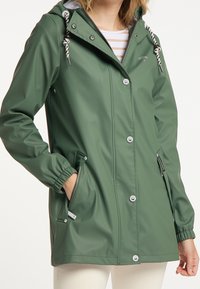 Femme portant une veste imperméable verte avec capuche, cordons rayés et poches zippées, associée à un pantalon blanc.