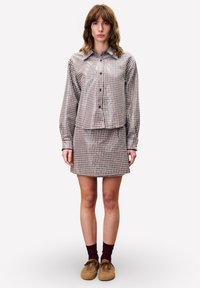 Vrouw staat recht vooruit kijkend, draagt een bruine geruite blouse met knopen, bijpassende rok, bordeauxrode sokken en bruine pluche schoenen.