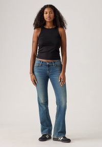 Femme aux cheveux bouclés portant un haut sans manches noir, un jean évasé bleu et des baskets noires se tient devant un fond clair uni.