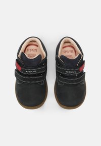 Scarpe nere in suede con due cinturini in Velcro, suole in gomma marrone e un accento rosso sul cinturino. L'interno è realizzato con materiale traspirante.