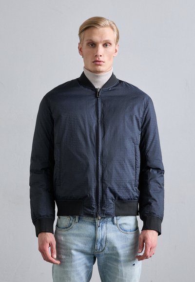 Veste de bombardier marine avec une surface texturée, fermeture éclair, col et poignets côtelés, associée à un jean léger bleu délavé.