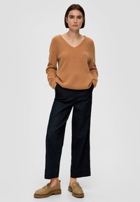 Sweter dzianinowy w serek w jasnobrązowym kolorze, zestawiony z szerokimi spodniami z ciemnego dżinsu oraz tanowymi zamszowymi mokasynami z ozdobnymi akcentami.