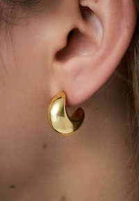 Boucle d'oreille en forme de créole de couleur dorée avec une finition lisse et polie. Le design présente une forme incurvée évoquant une onde qui épouse le lobe de l'oreille.