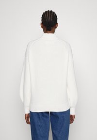Pull en tricot blanc avec un col haut, des accents côtelés et des manches larges. Porté sur un pantalon en denim bleu, mettant en avant un design texturé et douillet.