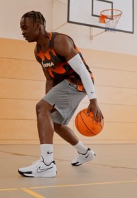 Joueur de basket-ball portant un maillot sans manches orange et noir à motifs, un short gris et des baskets blanches Nike, dribblant un ballon de basket à l'intérieur.