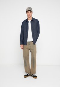 Μοντέλο που φοράει ένα μπλε σκούρο πουκάμισο με κουμπιά, λευκό μπλουζάκι, ανοιχτό καφέ παντελόνι με φαρδιές μπατζάκια και μαύρα αθλητικά παπούτσια με μπεζ λεπτομέρειες, ολοκληρωμένο με ένα γκρι καπέλο.