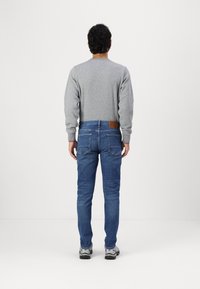Grauer Sweatshirt, blaue Slim-Fit-Jeans mit einem braunen Lederpatch und grauen Sneakern. Ansicht von hinten vor einem weißen Hintergrund.