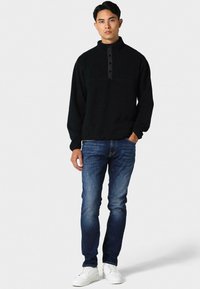 Juodas vilnos megztinis su stovimu apykakle ir sagų eilute, derinamas su mėlynais siaurais džinsais ir baltais sportbačiais.