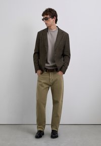 Hombre con blazer marrón, suéter beige, pantalones caqui, zapatos negros y gafas de sol oscuras de pie frente a una pared clara y sencilla.