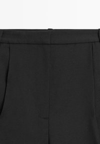 Pantalon noir avec une ceinture structurée, des plis à l'avant et une texture lisse. Design simple sans éléments visibles en métal.