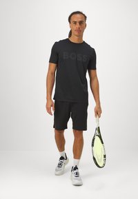 Camiseta deportiva de manga corta negra con el logo "BOSS", combinada con pantalones cortos negros y zapatillas deportivas blancas. Sosteniendo una raqueta de tenis.