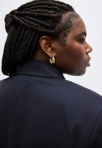 Profile view of a person with braided hair wearing gold hoop earrings and a dark navy blazer against a plain white background.