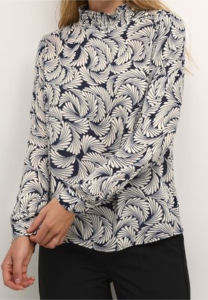 Femme portant un chemisier bleu marine à manches longues avec un motif abstrait blanc en forme d'éventail et un col à volants, ajustant sa manche, associé à un pantalon noir.