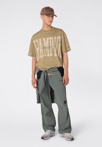 Joven con una gorra marrón, camiseta grande de Champion color beige, pantalones grises, zapatillas y una chaqueta atada alrededor de la cintura, de pie contra un fondo blanco.