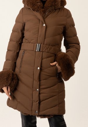 Brązowa puchowa kurtka z paskiem, zatrzaskami, kieszeniami na zamek i futrzanym wykończeniem na kołnierzu i mankietach, noszona na czarnych spodniach.