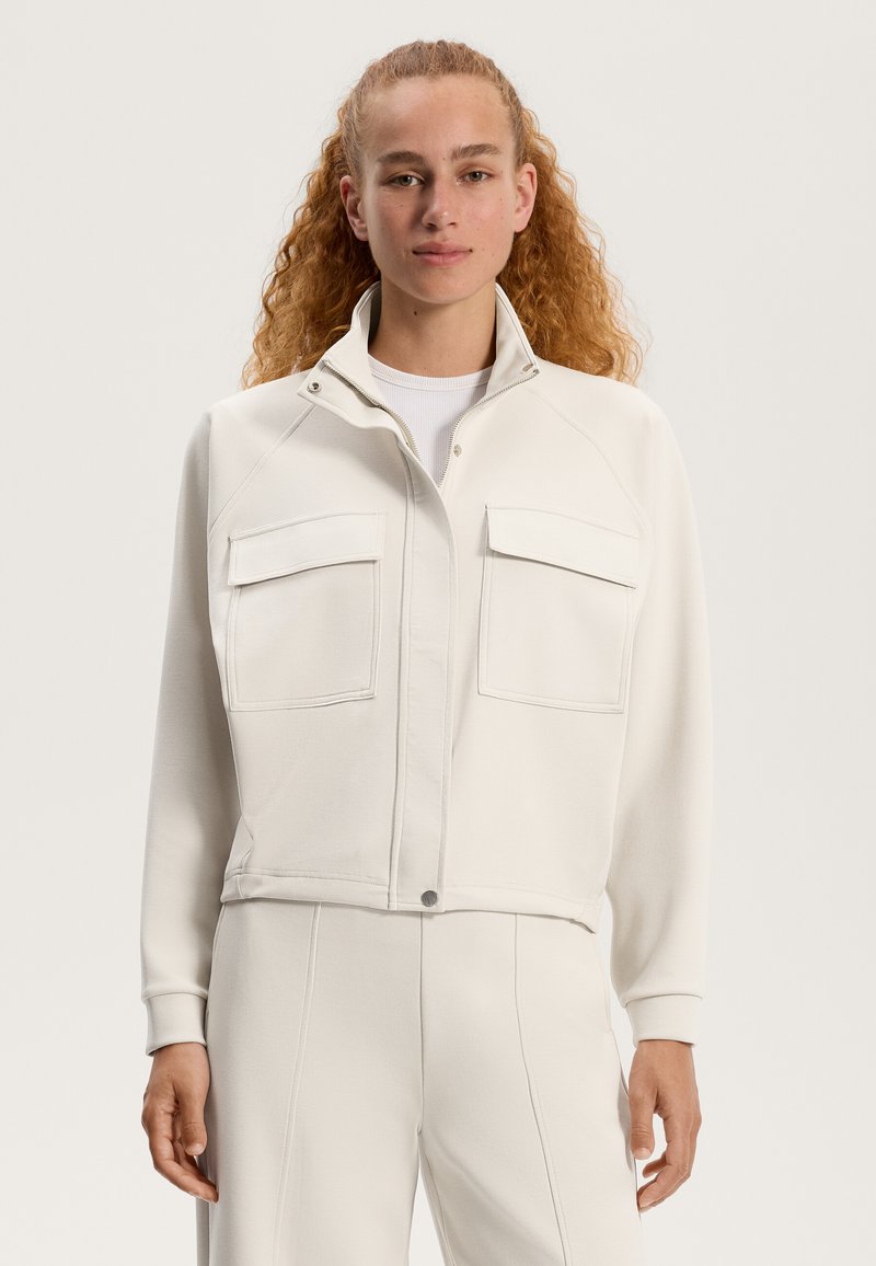 Weißes, kurzes Jackett aus weichem Stoff mit Reißverschluss, hohem Kragen und zwei Brusttaschen mit Klappen. Glatte Textur.