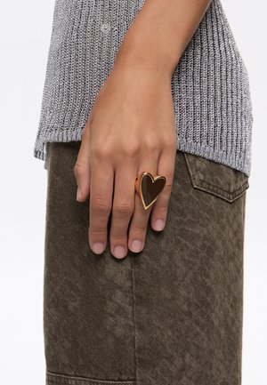 Anello a forma di cuore in oro con un centro in smalto marrone scuro. Mano visibile che indossa l'anello; sullo sfondo si vede un top a maglia grigia testurizzata e una gonna marrone.