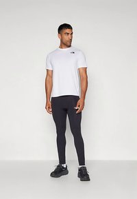 Homem vestindo uma camisa branca de manga curta da The North Face, leggings pretas justas e sapatos desportivos pretos, em pé contra um fundo simples.