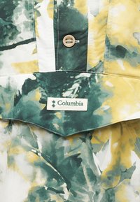Κοντινή λήψη υφάσματος με πράσινο, κίτρινο και λευκό σχέδιο, με ένα καφέ κουμπί και ετικέτα της Columbia Sportswear Company.