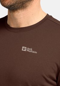 Mann mit Bart, der ein braunes Jack Wolfskin T-Shirt trägt, das Logo auf der Brust zeigt, beschnitten an Hals und Schultern.