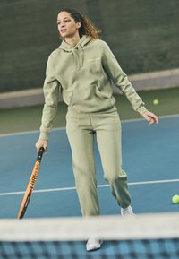 Casaco capuz verde claro e calças de treino combinando, feitos de um tecido suave. Apresenta um bolso na frente e um detalhe de logotipo discreto. O jogador segura uma raquete de ténis laranja.