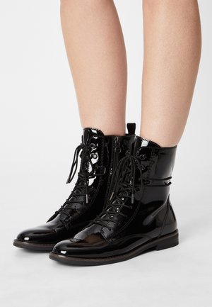 Bottines en cuir verni noir brillant à lacets avec fermeture éclair latérale portées sur des jambes nues, sur fond blanc.