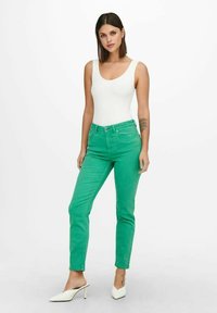 Green denim jeans with a slim fit, paired with a white bodysuit. The model wears white heeled mules, showcasing a simple and modern look.