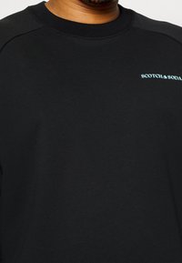 Felpa nera realizzata in morbido tessuto, con collo a giro e maniche raglan. Include il logo azzurro "SCOTCH & SODA" sul petto.