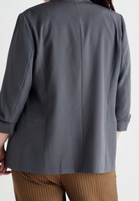 Šedý blazer s třičtvrtinovými rukávy, strukturovaný střih, hladká tkanina a jemné detaily šití. Kombinovaný s hnědými pruhovanými kalhotami.